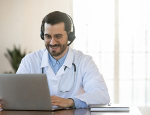 Smiling,Young,Doctor,In,Headset,Consulting,Patient,Online,,Using,Laptop,