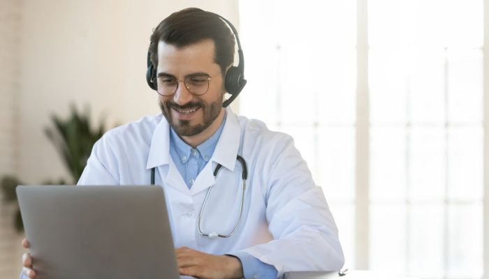 Smiling,Young,Doctor,In,Headset,Consulting,Patient,Online,,Using,Laptop,